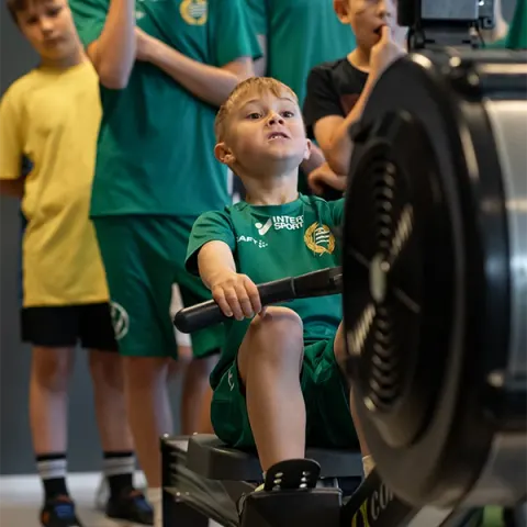 En pojke i fotbollströja drar i en roddmaskin medan lagkamraterna tittar på.
