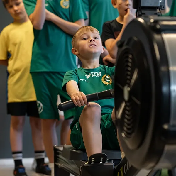 En pojke i fotbollströja drar i en roddmaskin medan lagkamraterna tittar på.