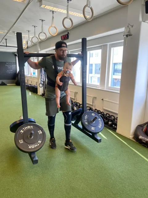 En man står i gymmet med ett litet barn i bärsele bredvid en skivstång.