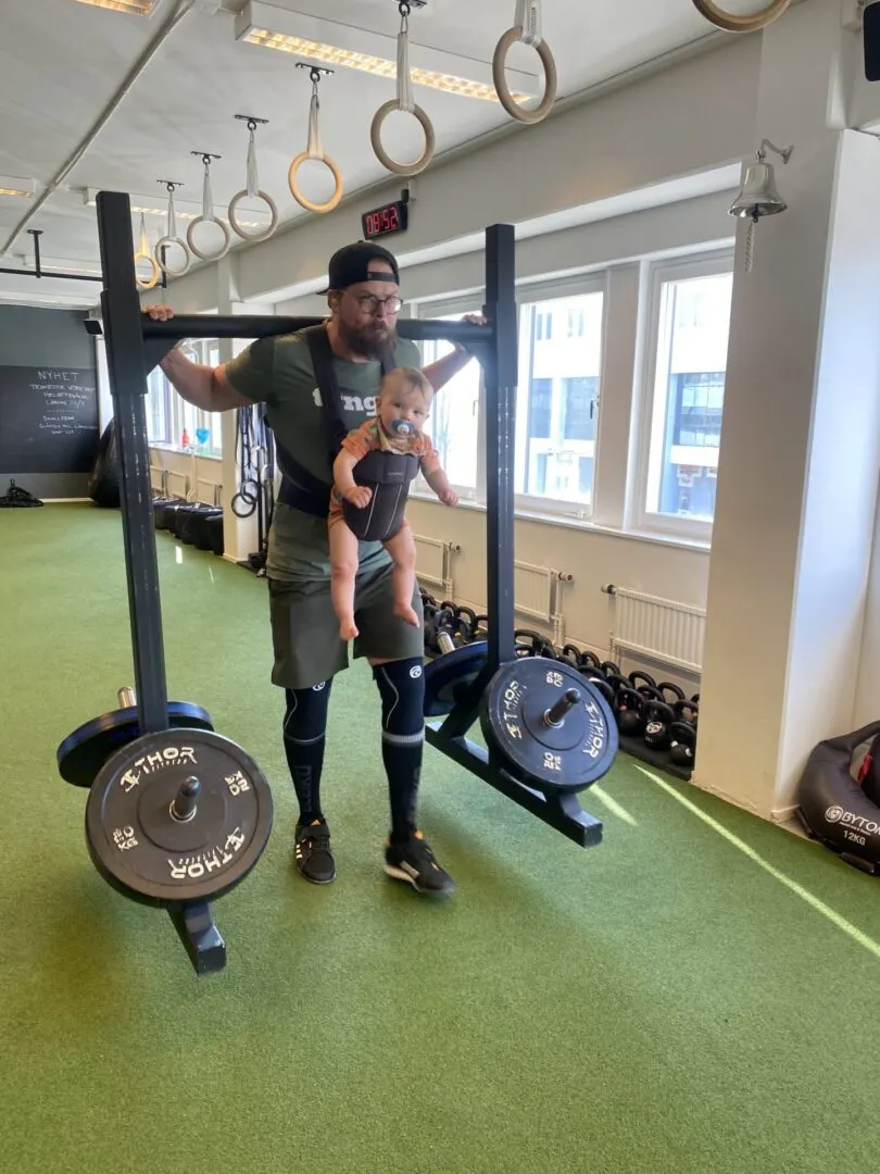 En man står i gymmet med ett litet barn i bärsele bredvid en skivstång.