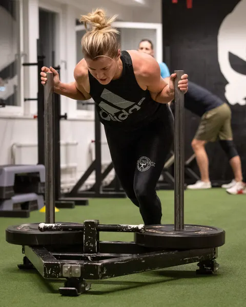 En kvinna trycker en tungt lastad släde över gymmets konstgräs.