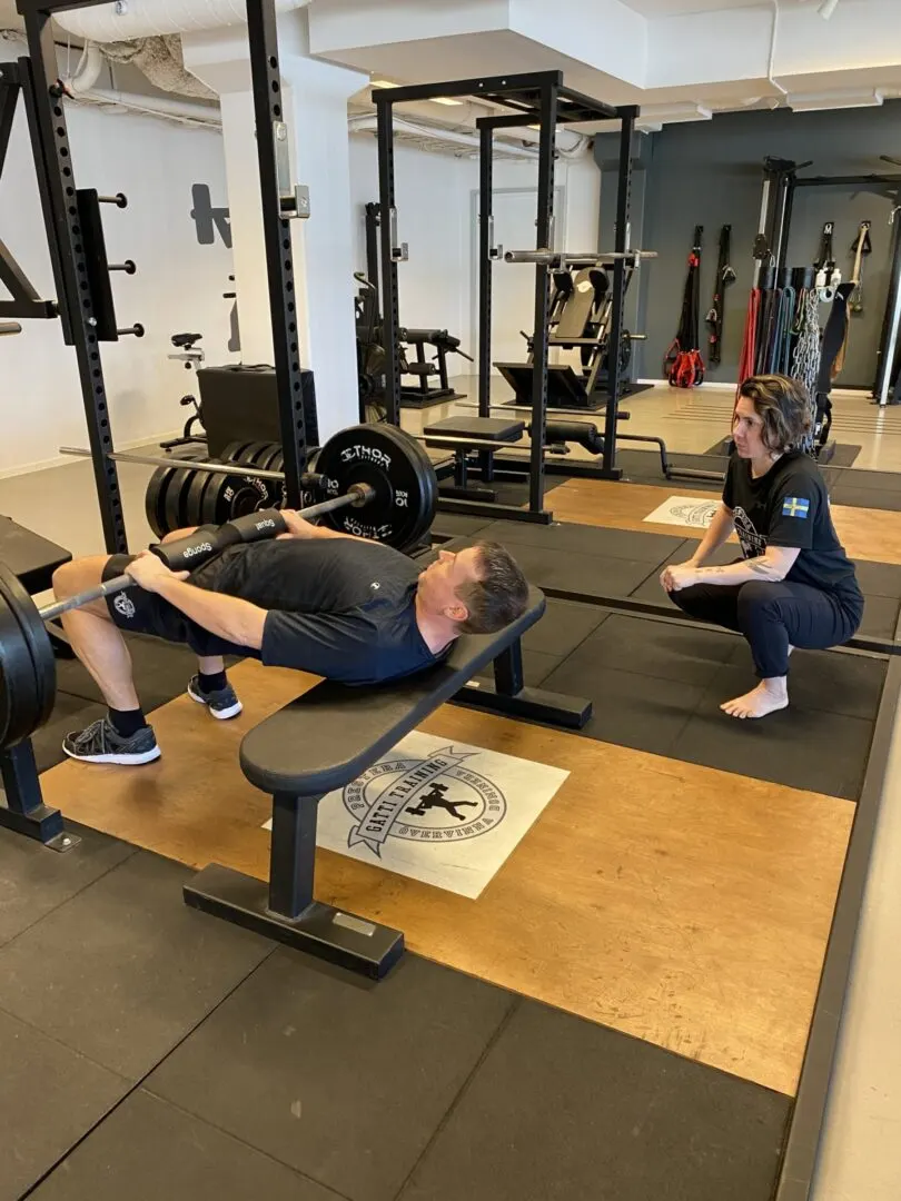 En man gör bänkpress medan en coach sitter på huk bredvid och följer lyftet.