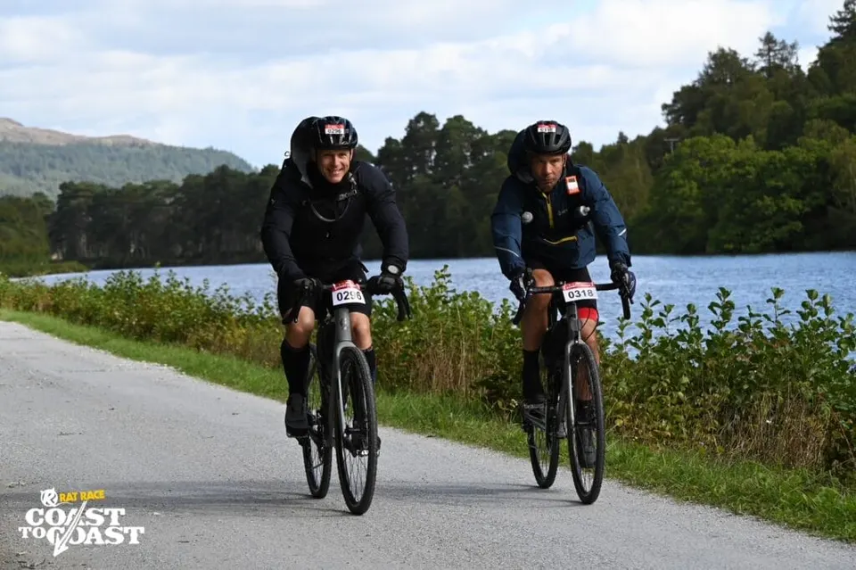 Två cyklister i hjälm cyklar längs en sjö i ett landsvägslopp.