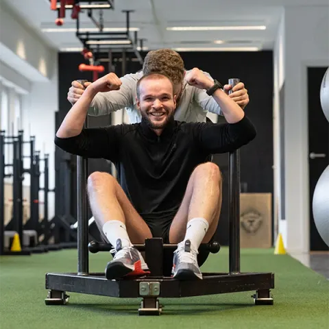 En leende man sitter i en slädmaskin medan en coach peppar honom bakifrån.