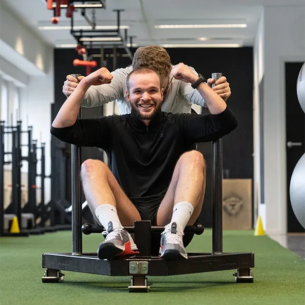 En leende man sitter i en slädmaskin medan en coach peppar honom bakifrån.