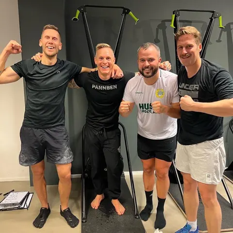 Fyra män i träningskläder poserar glatt och spänner armarna i gymmet.