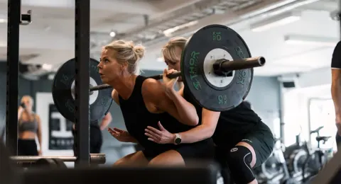 En kvinna gör knäböj med skivstång medan en träningskompis står redo bakom henne.