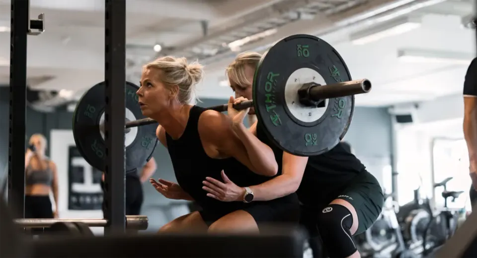 En kvinna gör knäböj med skivstång medan en träningskompis står redo bakom henne.