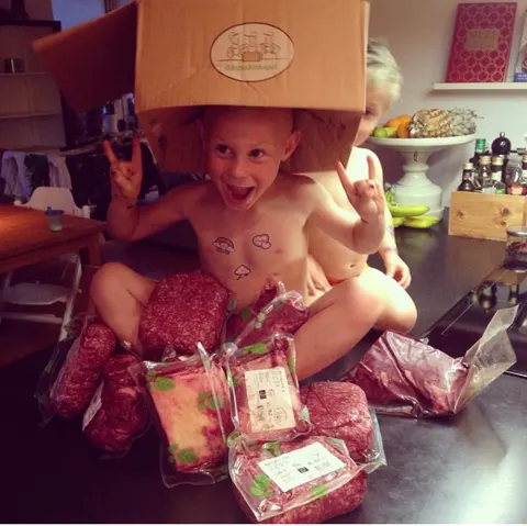Ett glatt barn med en kartong på huvudet sitter bland paket med köttfärs.
