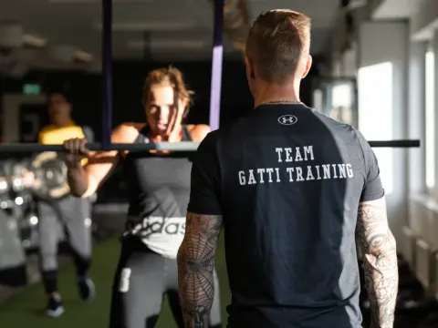 En coach i Team Gatti Training-tröja följer en kvinna som gör chins i en skivstångsställning.