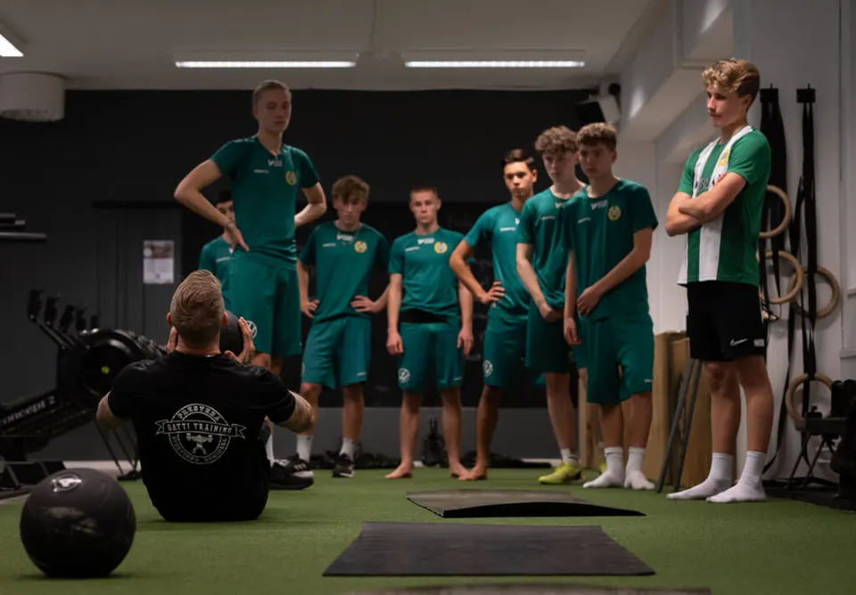 Unga fotbollsspelare i gröna kläder står i en halvcirkel och lyssnar på en coach.