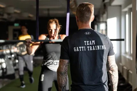 En coach i Team Gatti Training-tröja följer en kvinna som gör chins i en skivstångsställning.
