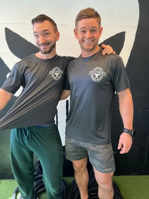 Två leende coacher i Gatti Training-tröjor står sida vid sida i gymmet.