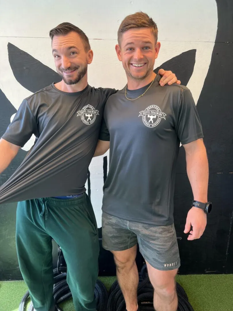 Två leende coacher i Gatti Training-tröjor står sida vid sida i gymmet.