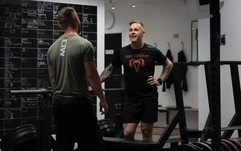 Två coacher samtalar framför svarta tavlan med träningsresultat i gymmet.