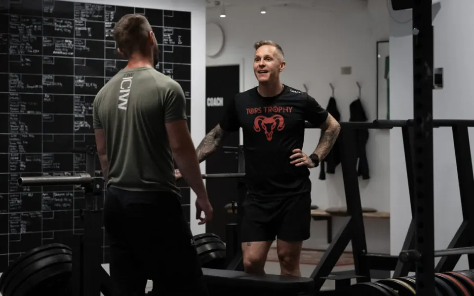 Två coacher samtalar framför svarta tavlan med träningsresultat i gymmet.