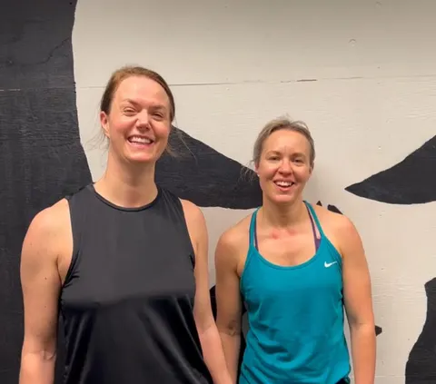 Två leende kvinnor i träningslinnen framför en målad vägg i gymmet.