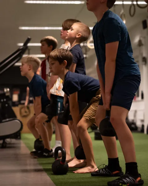 Barn står på rad och lyfter kettlebells med rak rygg under ett barnpass.