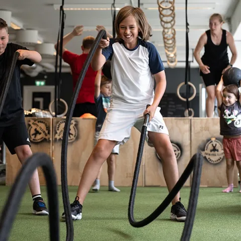 Barn drar i battle ropes på konstgräset medan en coach håller uppsikt.