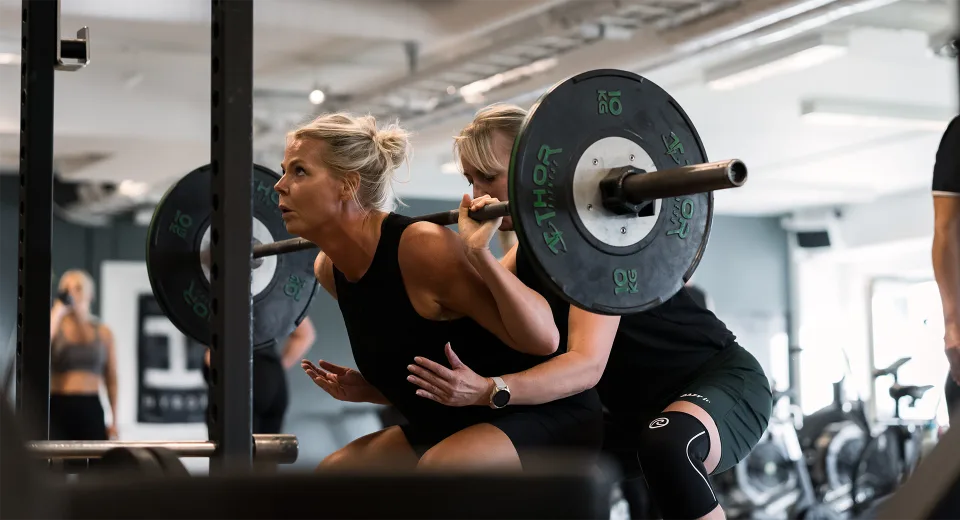 En medlem reser sig ur ett knäböj med skivstång medan en träningskompis står redo bakom henne.