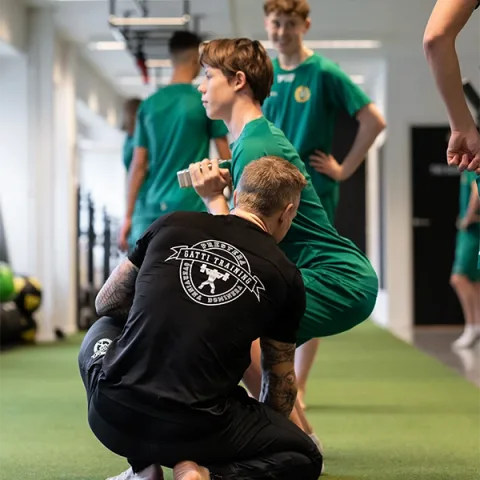 En coach sitter på huk och coachar unga spelare som tränar med hantlar.