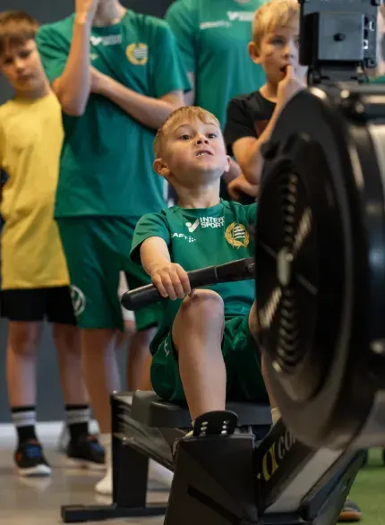 En pojke i fotbollströja drar i en roddmaskin medan lagkamraterna tittar på.